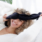 Woman wearing a black sleep mask with a white blanket and pillow in the background