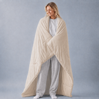 A woman wrapped in a large, quilted cream-colored comforter or duvet, standing against a solid gray studio background.