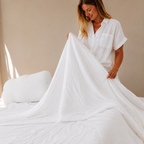 A woman in a white short-sleeve shirt holds up a large white duvet on a bed in a softly lit bedroom.