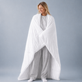 A woman wrapped in a large white quilted comforter or duvet, standing against a plain light gray studio background.