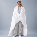 A woman wrapped in a large white quilted comforter or duvet, standing against a plain light gray studio background.