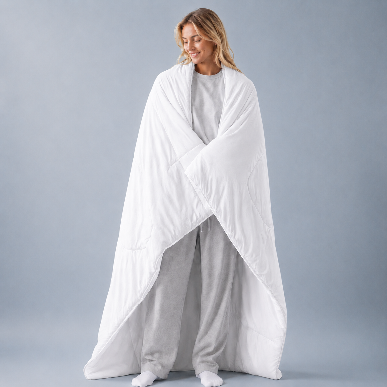 A woman wrapped in a large white quilted comforter or duvet, standing against a plain light gray studio background.
