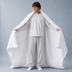 A man wearing a light gray pajama set stands against a gray studio background, holding open a large white quilted comforter or blanket draped over his shoulders like a cape.
